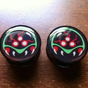 May include: A pair of black and green ear gauges featuring a printed design of a video game character. The character has three red eyes and a green shell.