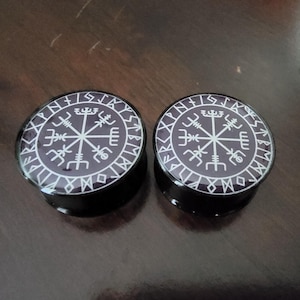 May include: Pair of black and white ear gauges with a compass design and runes around the edge.