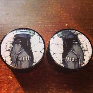 May include: Pair of black ear gauges featuring a detailed illustration of a titan's face breaking through a wall. The design is in shades of gray and white, with a black border. These gauges are likely made of a durable material.