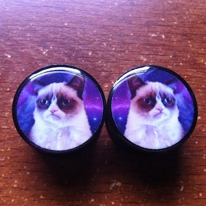 May include: A pair of black ear gauges with a galaxy background and a picture of a grumpy cat on each gauge.