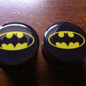 May include: Two black Batman logo plugs. Each plug features a yellow bat symbol within an oval on a black background. The plugs are round and appear to be made of a glossy material.