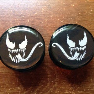 Puede incluir: Un par de dilatadores de oreja negros con un diseño de personaje Venom blanco. La cara del personaje se muestra con la boca abierta y una lengua larga.