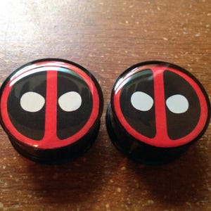 May include: A pair of black and red ear gauges with a white Deadpool logo.