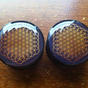 Könnte beinhalten: Zwei runde, schwarze Ohrstecker mit einem goldenen geometrischen Muster. Das Muster ist ein Lebensblume-Design.