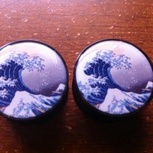Puede incluir: Un par de dilatadores de oreja negros y blancos con una imagen azul y blanca de una ola. La imagen recuerda la famosa estampa japonesa en madera, "La Gran Ola de Kanagawa".