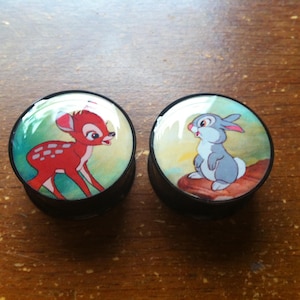 May include: A pair of black ear gauges with colorful images of Bambi and Thumper from Disney's Bambi. The Bambi gauge features a red deer with white spots on a green and yellow background. The Thumper gauge features a gray rabbit with a pink nose on a blue and green background.