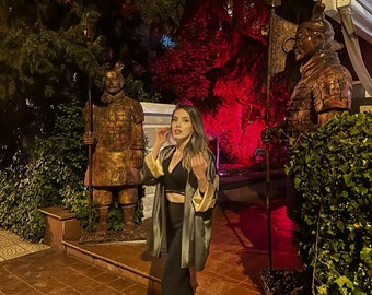 Puede incluir: Una mujer con una chaqueta verde y dorada se encuentra frente a dos estatuas de guerreros. Las estatuas son de bronce y llevan armadura tradicional. La mujer sonríe y mira a la cámara.