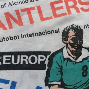 May include: White t-shirt with a black and white graphic print of a soccer player wearing a green jersey with the number 8. The text "Jogo de futobol Internacional" and "EUROP" are also printed on the shirt.
