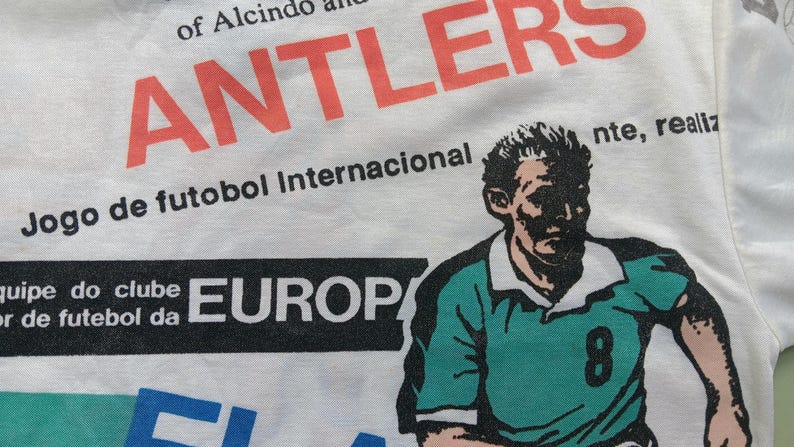 May include: White t-shirt with a black and white graphic print of a soccer player wearing a green jersey with the number 8. The text "Jogo de futobol Internacional" and "EUROP" are also printed on the shirt.