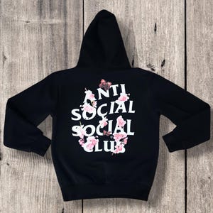 May include: Black hooded sweatshirt with the text "ANTI SOCIAL SOCIAL CLUB" in white, adorned with pink floral and butterfly accents. The hoodie has a drawstring hood and long sleeves.