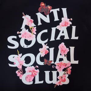 May include: Black sweatshirt with the words "ANTI SOCIAL SOCIAL CLUB" in white block letters. Pink cherry blossoms and butterflies surround the text. The sweatshirt has long sleeves.