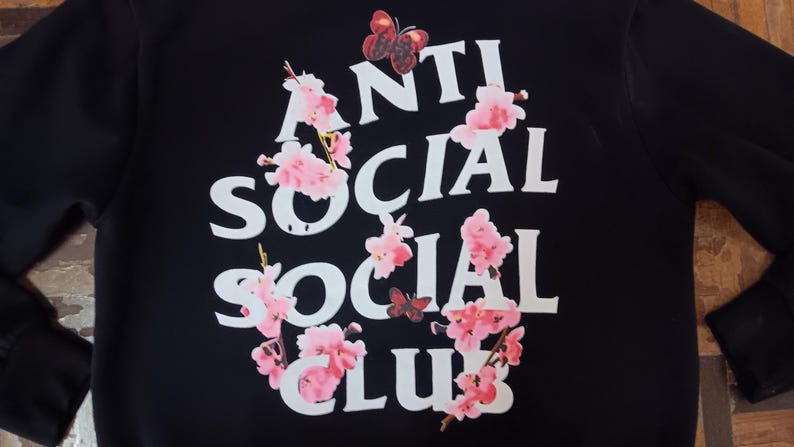 May include: Black sweatshirt with the words "ANTI SOCIAL SOCIAL CLUB" in white block letters. Pink cherry blossoms and butterflies surround the text. The sweatshirt has long sleeves.