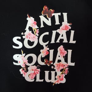 May include: Black garment featuring the words "ANTI SOCIAL SOCIAL CLUB" in white, adorned with pink floral and butterfly graphics. The design is centered and prominent against the dark background.