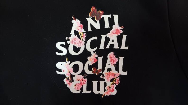 May include: Black garment featuring the words "ANTI SOCIAL SOCIAL CLUB" in white, adorned with pink floral and butterfly graphics. The design is centered and prominent against the dark background.