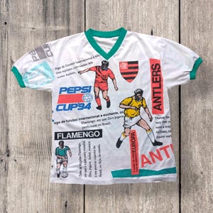 May include: White soccer jersey with a green collar and sleeves. The jersey has a variety of colorful graphics, including the Pepsi Cup '94 logo, the Flamengo logo, and the Antlers logo. The text "Jogo de futobol Internacional e excitante, com equipes convidadas tais como Antlers" is printed on the jersey.