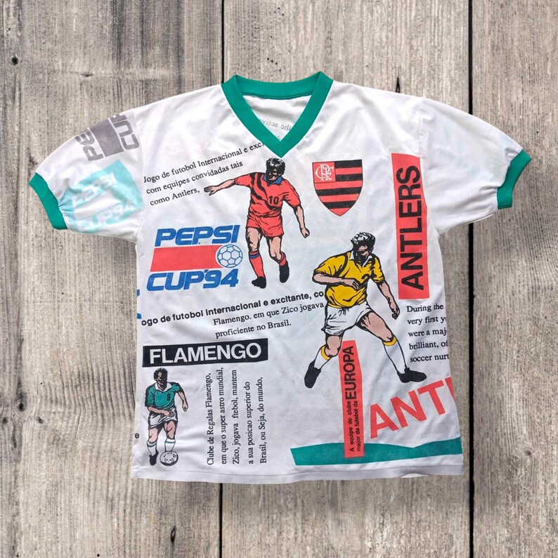 May include: White soccer jersey with a green collar and sleeves. The jersey has a variety of colorful graphics, including the Pepsi Cup '94 logo, the Flamengo logo, and the Antlers logo. The text "Jogo de futobol Internacional e excitante, com equipes convidadas tais como Antlers" is printed on the jersey.