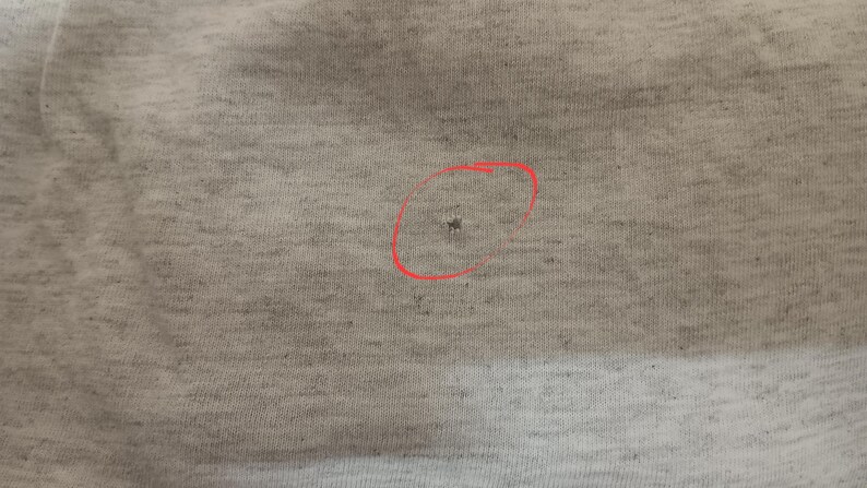 May include: Close-up of a light gray fabric with a small hole circled in red. The fabric appears to be a knit material, with a slightly textured surface. The hole is small and dark, contrasting with the lighter background.