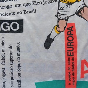 May include: White t-shirt with a red and black graphic of a soccer player. The graphic includes the text "EUROPA" and "A equipe do clube major de futebol da". The shirt also has the text "LAMENGO" and "Clube de Regalas Flamengo" in black.