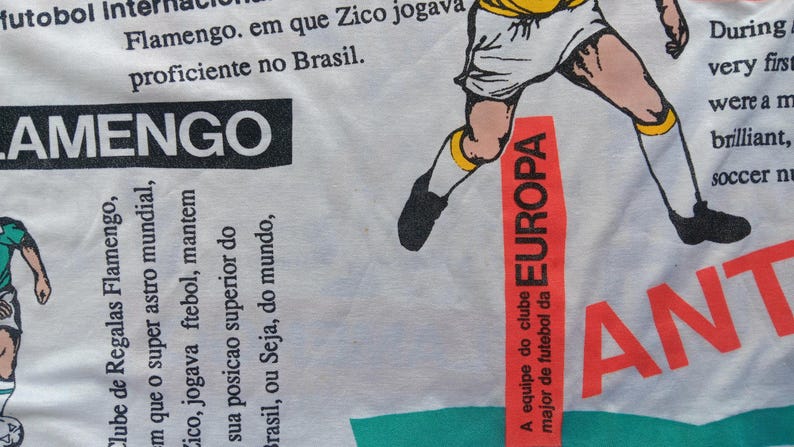 May include: White t-shirt with a red and black graphic of a soccer player. The graphic includes the text "EUROPA" and "A equipe do clube major de futebol da". The shirt also has the text "LAMENGO" and "Clube de Regalas Flamengo" in black.