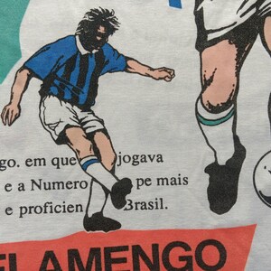 May include: A white t-shirt with a red, white, and blue graphic of a soccer player. The text "PEPSI" is printed on the left side of the shirt. The text "FLAMENGO" is printed in red at the bottom of the shirt. The text "Flamengo. em que jogava futebol, e a Numero pe mais popular e proficien Brasil." is printed in black on the shirt.