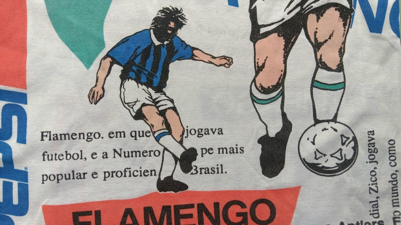May include: A white t-shirt with a red, white, and blue graphic of a soccer player. The text "PEPSI" is printed on the left side of the shirt. The text "FLAMENGO" is printed in red at the bottom of the shirt. The text "Flamengo. em que jogava futebol, e a Numero pe mais popular e proficien Brasil." is printed in black on the shirt.