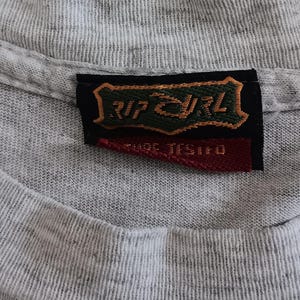 May include: Close-up of a heather gray t-shirt with a V-neck collar. A black and red tag with the text "RIP CURL" and "CARE TESTED" is sewn into the collar. The shirt's fabric has a textured appearance.