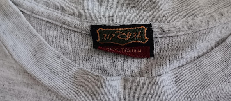 May include: Close-up of a heather gray t-shirt with a V-neck collar. A black and red tag with the text "RIP CURL" and "CARE TESTED" is sewn into the collar. The shirt's fabric has a textured appearance.
