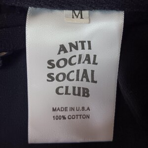 May include: Close-up of a white clothing tag with the text "ANTI SOCIAL SOCIAL CLUB" in bold, black letters. The tag also reads "MADE IN U.S.A" and "100% COTTON". A size "M" is printed at the top of the tag.