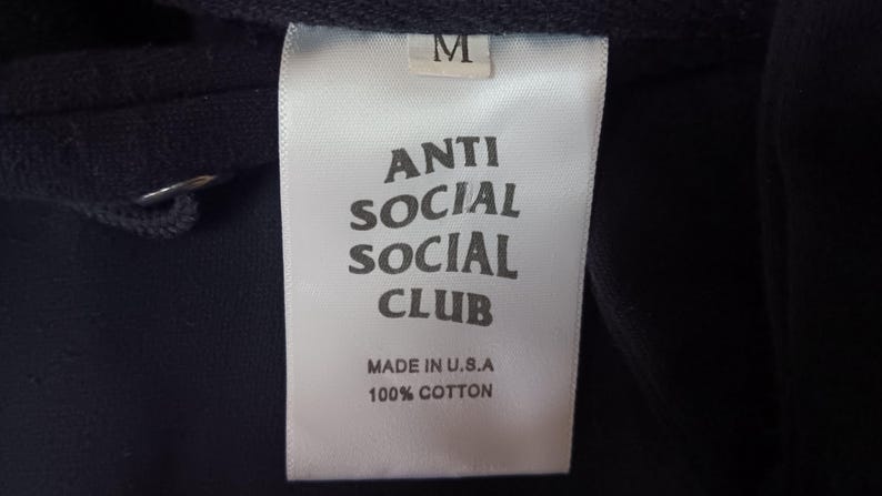May include: Close-up of a white clothing tag with the text "ANTI SOCIAL SOCIAL CLUB" in bold, black letters. The tag also reads "MADE IN U.S.A" and "100% COTTON". A size "M" is printed at the top of the tag.