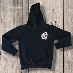 May include: Black hooded sweatshirt with a front pocket and drawstring hood. The sweatshirt features the text "ANTI SOCIAL SOCIAL CLUB" in white on the front. The garment is displayed on a wooden surface.