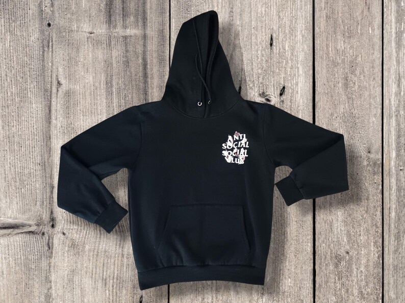 May include: Black hooded sweatshirt with a front pocket and drawstring hood. The sweatshirt features the text "ANTI SOCIAL SOCIAL CLUB" in white on the front. The garment is displayed on a wooden surface.