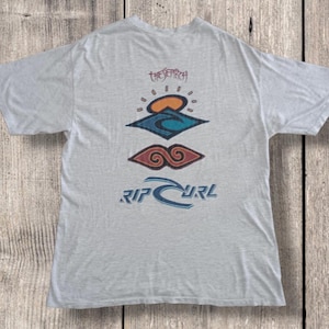 May include: Light gray t-shirt with a colorful graphic design. The design includes a sun, waves, and the text "RIP CURL" in blue. The shirt also features the text "THE SEARCH" in a script font.