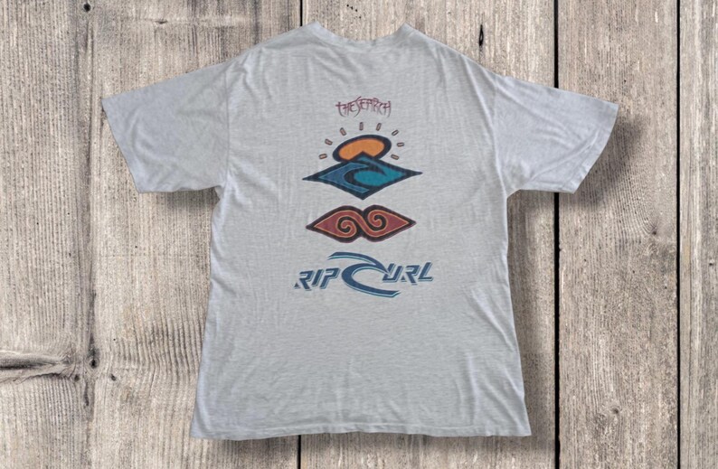 May include: Light gray t-shirt with a colorful graphic design. The design includes a sun, waves, and the text "RIP CURL" in blue. The shirt also features the text "THE SEARCH" in a script font.