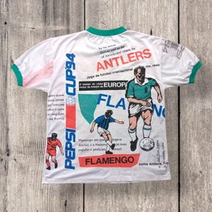 May include: White t-shirt with a green and pink trim. The shirt features a large graphic print of a soccer player in a green and white uniform with the text "Flamengo" and "Pepsi Cup 94".