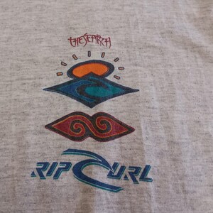 May include: Light gray t-shirt featuring a colorful graphic design. The design includes the text "THE SEARCH" above a stylized sun and wave motif, and the brand name "RIP CURL" in blue.
