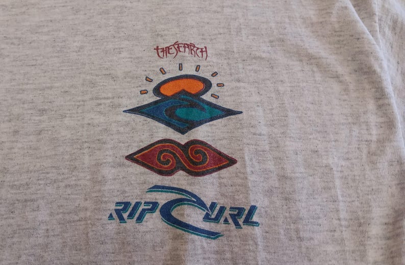 May include: Light gray t-shirt featuring a colorful graphic design. The design includes the text "THE SEARCH" above a stylized sun and wave motif, and the brand name "RIP CURL" in blue.