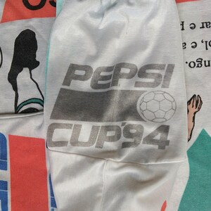 May include: A white t-shirt with a Pepsi Cup '94 logo featuring a soccer ball. The logo is in gray and white with the text "Pepsi Cup '94".