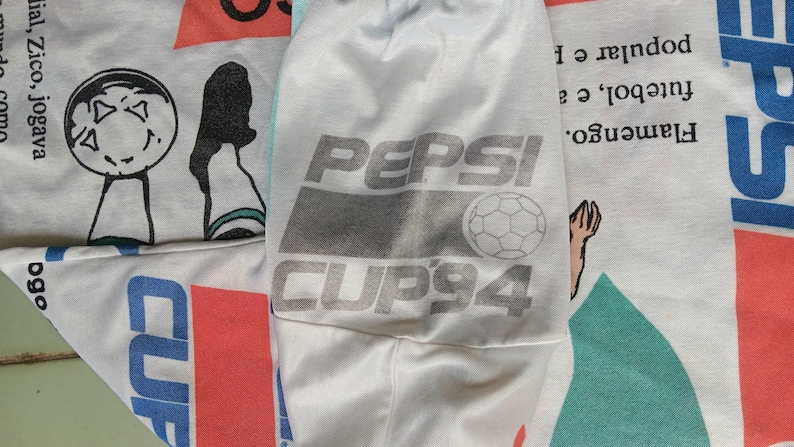 May include: A white t-shirt with a Pepsi Cup '94 logo featuring a soccer ball. The logo is in gray and white with the text "Pepsi Cup '94".