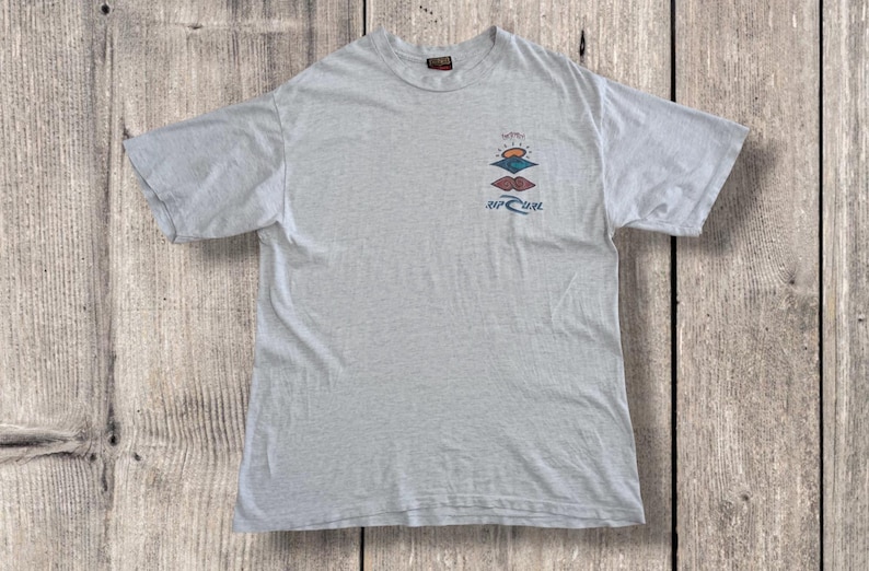May include: Light gray t-shirt with a crew neck and short sleeves. The shirt features a colorful graphic of a sunset and the text "Rip Curl". The shirt is made of a soft, comfortable material.