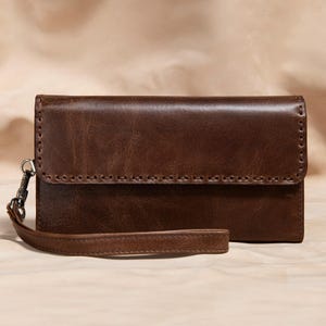May include: A brown leather wallet with a wrist strap. The wallet has a rectangular shape with a flap closure and visible stitching along the edges. The leather has a textured appearance and a metal clasp on the strap.