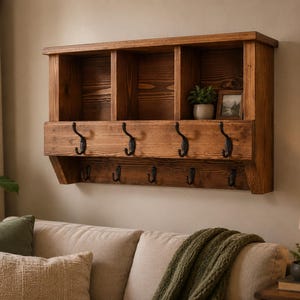 May include: A wall-mounted wooden shelf unit featuring three open storage compartments on top and two rows of black metal hooks. The unit is crafted from stained wood with a visible grain pattern and includes a small potted plant and a framed picture.