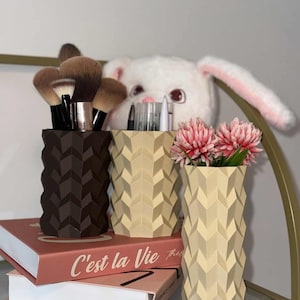 May include: Three geometric patterned containers in brown, beige, and cream. The brown container holds makeup brushes, the beige one holds pens, and the cream one holds pink flowers. The containers sit on a shelf with books.