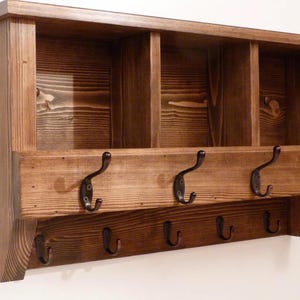 May include: A rustic wooden wall-mounted organizer with a dark brown finish. It features three cubby shelves at the top, a row of five metal hooks below, and a lower row of hooks. The wood grain is visible, adding to its natural aesthetic.