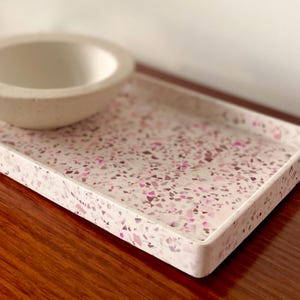 May include: A rectangular tray and a circular bowl featuring a white base with embedded multi-tonal pink and purple terrazzo chips. The tray has raised edges and a flat surface, while the bowl has a smooth, curved interior and exterior finish.