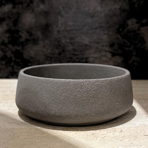 May include: A circular, low-profile bowl with a dark grey, textured, matte finish. The vessel features thick, solid walls and a wide, open top, presenting a uniform, granular surface texture throughout the exterior and interior rim.