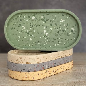 May include: A stack of four oval-shaped trays with a terrazzo pattern. The top tray is sage green with white and dark green flecks. The lower trays are beige, grey, and yellow, each featuring multi-coloured stone-like fragments embedded throughout.