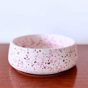 May include: A round, shallow bowl featuring a white base with a speckled terrazzo pattern in various shades of pink, magenta, and grey. The bowl has a smooth, matte finish and a wide, open top, showcasing a consistent confetti-like design.