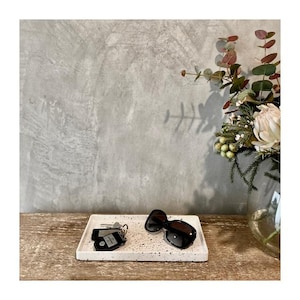May include: A rectangular, speckled white tray holds a set of keys and black sunglasses. The tray sits on a wooden surface, with a grey wall in the background. A vase of flowers and greenery is on the right.