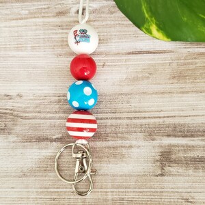 May include: A red, white, and blue beaded keychain with a red, white, and blue patterned bead, a blue bead with white polka dots, a red bead, and a white bead with a graphic of a person in a red and white striped hat. The keychain has a silver metal clasp.