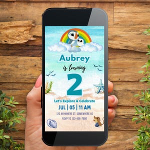 Puede incluir: Un teléfono inteligente muestra una colorida invitación para el segundo cumpleaños de Aubrey. El diseño presenta un arcoíris, frailecillos de dibujos animados e ilustraciones con temática de playa. El texto incluye el nombre "Aubrey", el número "2" y detalles del evento.